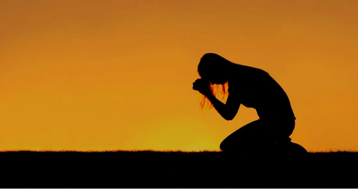 A person kneeling in prayer against a glowing sunset sky